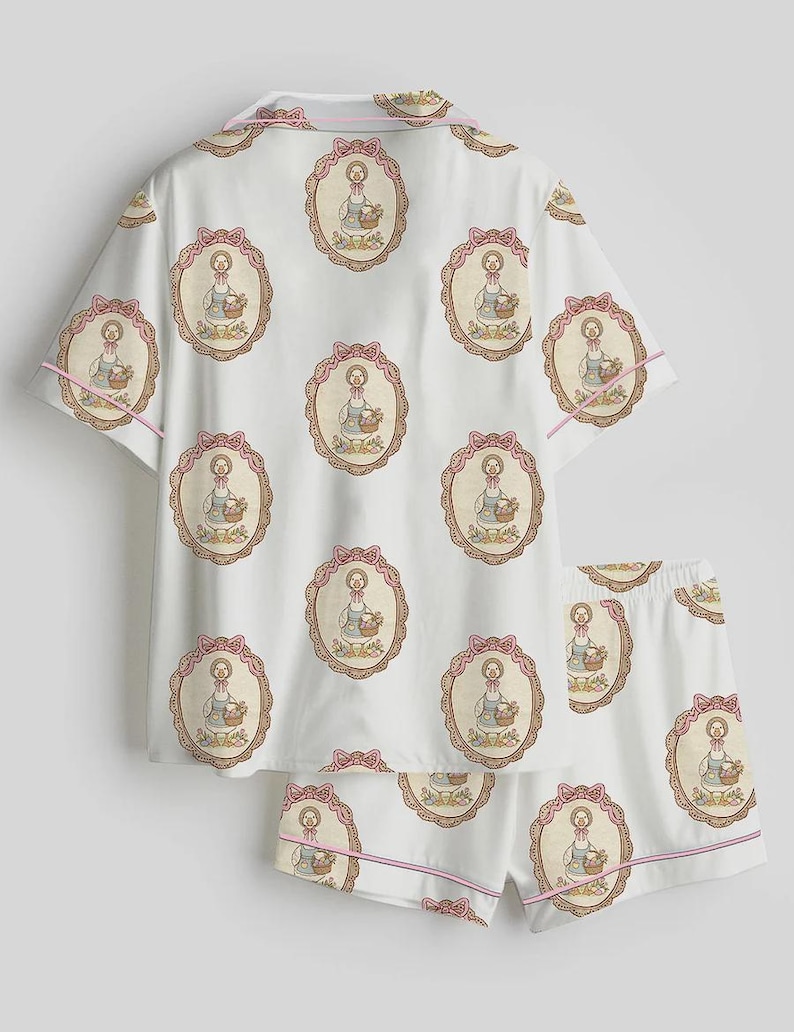 Może przedstawiać: Biały zestaw piżamowy z powtarzającym się wzorem ilustracji gęsi w owalnej ramie, z r&oacute;żowymi kokardkami i wykończeniami. Zestaw zawiera top z kr&oacute;tkim rękawem i pasujące szorty.