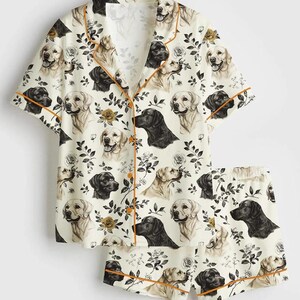 Pu&ograve; includere: Pigiama color crema con un motivo ripetuto di Labrador neri, marroni e crema, insieme ad accenti floreali. La maglia a maniche corte e i pantaloncini hanno bordi arancioni.