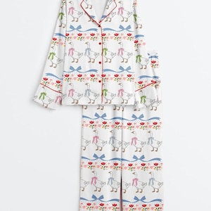Może przedstawiać: Biały zestaw piżamowy z powtarzającym się wzorem gęsi w kokardkach, czerwonymi wykończeniami i niebieskimi kokardami. Zestaw zawiera koszulę zapinaną na guziki i pasujące spodnie. Kwiatowe i sercowe akcenty dopełniają projekt.