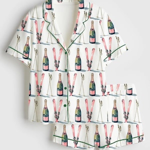 Puede incluir: Conjunto de pijama blanco con un estampado repetido de botellas de champán, esquís rosas y bastones de esquí. La parte superior de manga corta y los pantalones cortos están ribeteados con ribetes verdes. El conjunto está hecho de una tela suave y ligera.