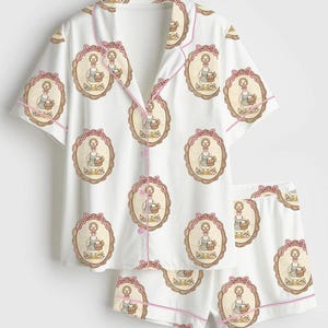 Może przedstawiać: Biały zestaw piżamowy z powtarzającym się wzorem gęsi niosącej kosz, oprawionej w owal z r&oacute;żową kokardą. Zestaw zawiera koszulę zapinaną na guziki z kr&oacute;tkim rękawem i pasujące szorty, wykończone r&oacute;żową lam&oacute;wką.