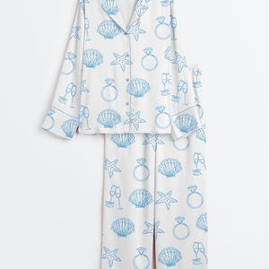 Op de afbeelding: Witte pyjamaset met een blauw patroon van zeeschelpen, zeesterren, champagneglazen en diamanten ringen. De top met lange mouwen heeft een knoopsluiting en een reverskraag. De bijpassende broek heeft een relaxte pasvorm.