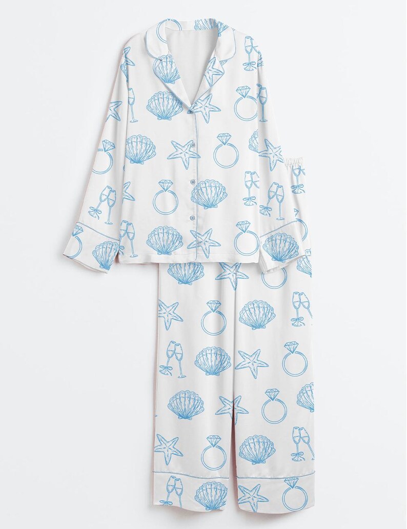 Op de afbeelding: Witte pyjamaset met een blauw patroon van zeeschelpen, zeesterren, champagneglazen en diamanten ringen. De top met lange mouwen heeft een knoopsluiting en een reverskraag. De bijpassende broek heeft een relaxte pasvorm.