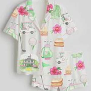 May include: White pajama set with a golf-themed pattern. The set includes a short-sleeved button-down shirt and matching shorts. The pattern features golf carts, golf balls, pink flowers, and a golf course scene.