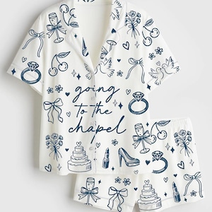 May include: White pajama set with a button-down shirt and shorts, adorned with navy blue illustrations of wedding-themed elements. The shirt features the text "going to the chapel". The set includes drawings of rings, cakes, and champagne glasses.