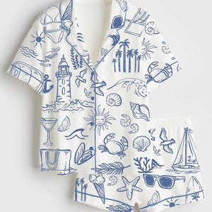 May include: White pajama set with a blue beach-themed print. The short-sleeved top and shorts feature illustrations of a lighthouse, seashells, crabs, and sunglasses. The set has blue trim.