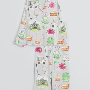May include: Light gray pajama set with a golf-themed pattern. The set features a button-down top and pants, adorned with golf carts, golf balls, golf clubs, sandwiches, pink flowers, and a green golf course. The trim is pink.