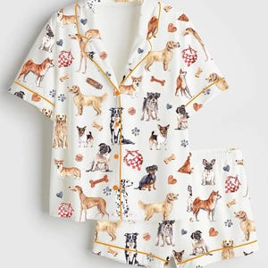 Peut inclure: Ensemble de pyjama blanc avec un motif de chiens. Le haut à manches courtes et le short présentent diverses races de chiens, des empreintes de pattes, des cœurs et des os. L'ensemble est bordé d'un passepoil doré.