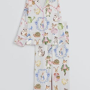 May include: Light gray pajama set with a whimsical print of woodland creatures, including rabbits, foxes, and owls. The long-sleeved top and pants feature pink piping and a button-down front. The pajama set is made of a soft, lightweight fabric.