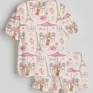 K&ouml;nnte beinhalten: Ein zweiteiliger Schlafanzug mit einem sich wiederholenden Muster von Pariser Caf&eacute;szene. Das Design umfasst den Eiffelturm, Caf&eacute;tische mit Sonnenschirmen, Geigen und die Texte "Paris" und "Bon Jour" in Rosa und Beige.