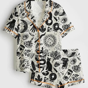 May include: White pajama set with a black celestial print featuring moons, suns, snakes, and hands. The short-sleeved top and shorts have orange trim. The set includes a button-down shirt and shorts.