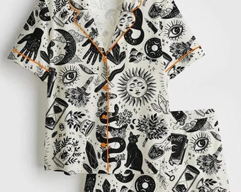 May include: White pajama set with a black celestial print featuring moons, suns, snakes, and hands. The short-sleeved top and shorts have orange trim. The set includes a button-down shirt and shorts.