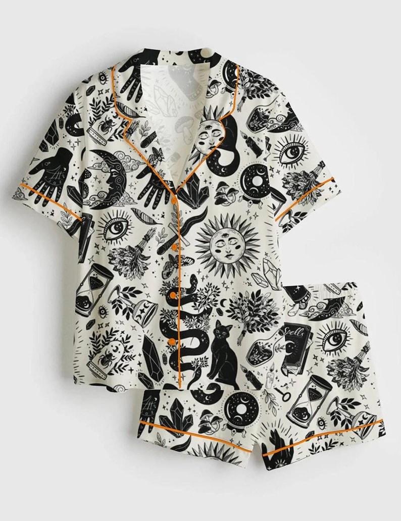 May include: White pajama set with a black celestial print featuring moons, suns, snakes, and hands. The short-sleeved top and shorts have orange trim. The set includes a button-down shirt and shorts.