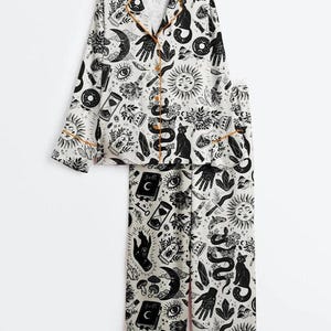 May include: White pajama set with a black celestial print featuring moons, suns, cats, and hands. The long-sleeved top and pants have orange trim. The button-down top has a notched collar.