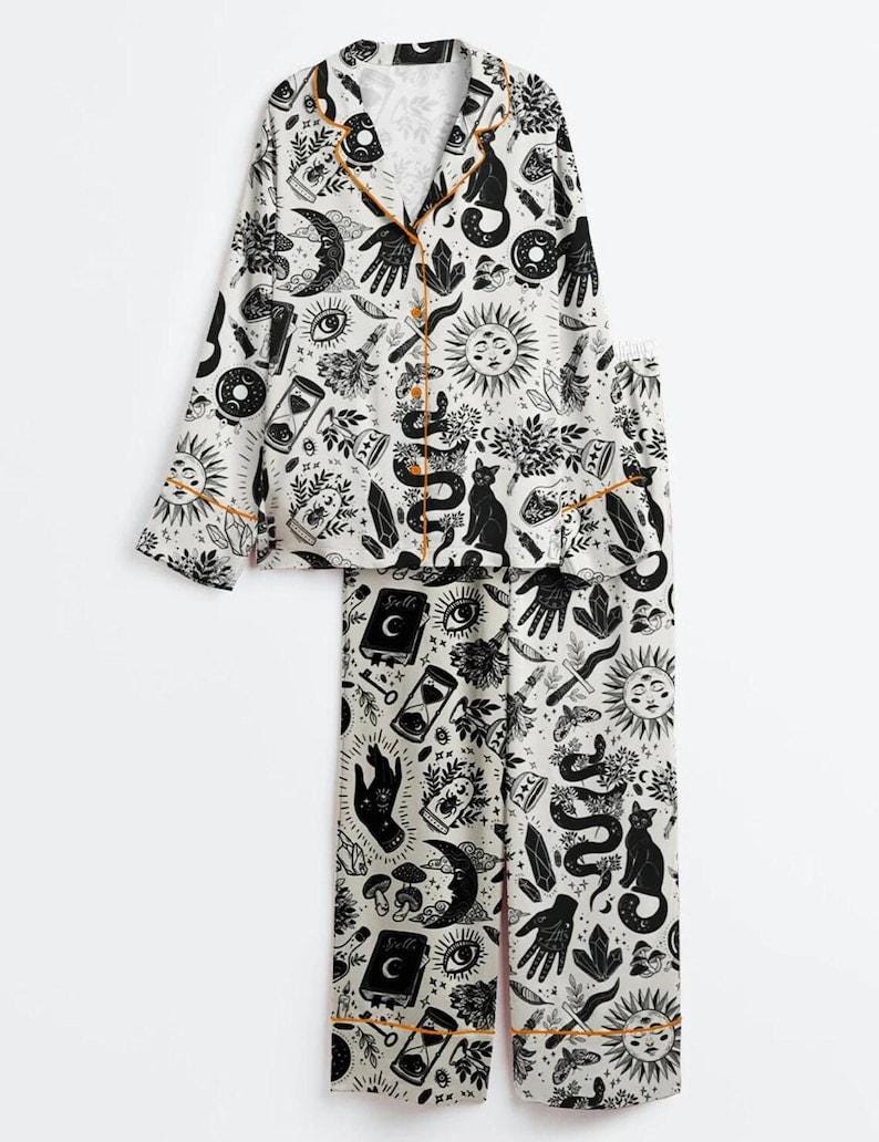 May include: White pajama set with a black celestial print featuring moons, suns, cats, and hands. The long-sleeved top and pants have orange trim. The button-down top has a notched collar.