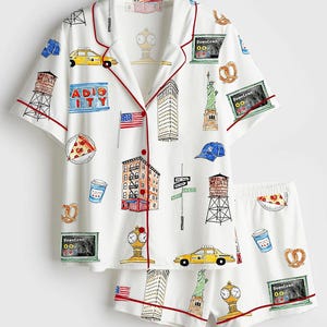 Pode incluir: Conjunto de pijama branco com tema de Nova York. A parte superior de manga curta e os calções apresentam acabamentos vermelhos e são cobertos por ilustrações coloridas de marcos, um táxi amarelo e alimentos. O conjunto inclui uma camisa abotoada e calções.