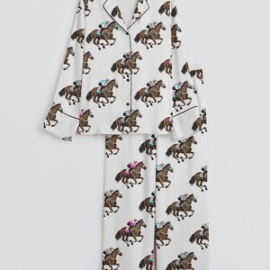 K&ouml;nnte beinhalten: Wei&szlig;es Pyjama-Set mit einem sich wiederholenden Muster aus braunen Rennpferden und Jockeys. Das lang&auml;rmlige Oberteil hat schwarze Paspeln und Kn&ouml;pfe. Die passende Hose hat ebenfalls schwarze Paspeln.