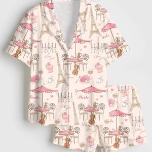 K&ouml;nnte beinhalten: Ein zweiteiliger hellrosa Pyjama-Set mit einem sich wiederholenden Muster von Pariser Caf&eacute;szene. Das Design umfasst den Eiffelturm, Tische mit Sonnenschirmen, Geigen und Cupcakes. Das Set ist mit Rosa, Braun und Wei&szlig; akzentuiert.