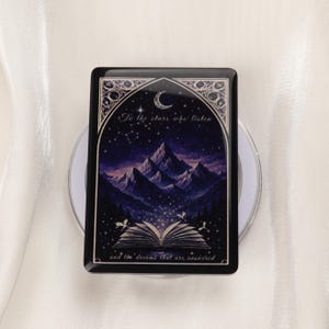 May include: A rectangular black resin coaster with a celestial design. The coaster features a mountain landscape under a starry night sky, a crescent moon, and the text "To the stars who listen and the dreams that are answered."