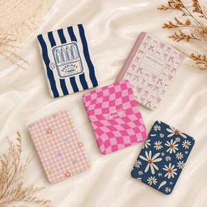 May include: A collection of small notebooks with various designs. One notebook features a sardine can illustration on a blue and white striped background. Other notebooks have pink checkered patterns, daisy patterns, and bow patterns. One notebook has the text "my weekend is booked".