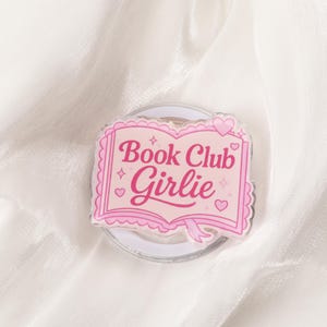 Op de afbeelding: Een roze en witte Book Club Girlie telefoonhouder. De houder heeft een roze open boekontwerp met de woorden "Book Club Girlie" in roze script. Het ontwerp bevat harten en sterren.