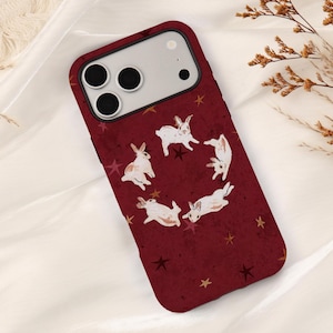 May include: A dark red smartphone case featuring a pattern of white rabbits arranged in a circular formation, interspersed with small gold and dark red stars. The case has a rectangular cutout for a triple-lens camera system on the upper left.