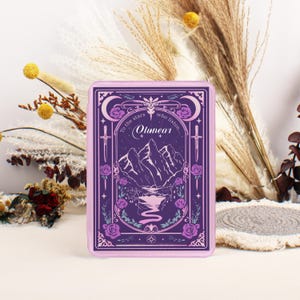 Puede incluir: Una funda de teléfono rectangular de color lavanda con una ilustración detallada. El diseño incluye una cordillera, un río, rosas y el texto "To the stars who listen... Olunear". Dos espadas y medias lunas enmarcan el diseño.