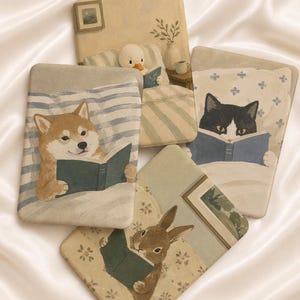 Op de afbeelding: Vier rechthoekige e-readerhoezen met illustraties van een Shiba Inu, een eend, een zwart-witte kat en een konijn, die elk in bed een boek lezen. De ontwerpen gebruiken zachte, gedempte tinten en tonen gezellige scènes met kussens en dekens.