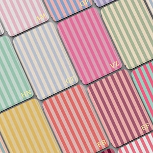 Puede incluir: Una colección de estuches rectangulares con rayas verticales en varios colores pastel y vivos. Cada estuche está personalizado con dos letras mayúsculas doradas en la esquina inferior, incluyendo iniciales como AM, OP, VZ, GT, HN y FB.