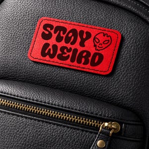 May include: Black backpack with a red patch that reads "STAY WEIRD" in a retro font. The patch features a small alien head graphic. The backpack has a zippered front pocket with a gold-colored zipper and pull.