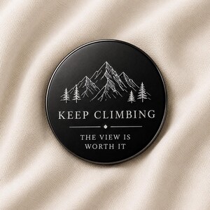 May include: A circular black pin featuring a white line art illustration of a mountain range and pine trees. The text reads KEEP CLIMBING above a horizontal line with a small diamond shape, followed by the text THE VIEW IS WORTH IT below.