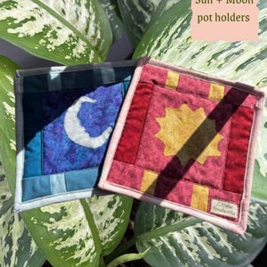 Könnte beinhalten: Zwei quadratische Topflappen mit Himmelsmotiven. Einer zeigt einen weißen Halbmond auf blauem Grund, der andere eine gelbe Sonne auf rotem Grund. Der Text "Sun + Moon pot holders" ist sichtbar. Die Topflappen befinden sich auf einem Hintergrund aus grünen Blättern.