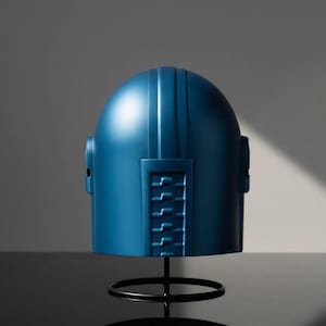 Puede incluir: Un casco azul con una parte superior redondeada y l&iacute;neas verticales, montado sobre un soporte de metal negro. El casco tiene un dise&ntilde;o rectangular en el lateral con una serie de l&iacute;neas horizontales. El fondo es gris neutro.