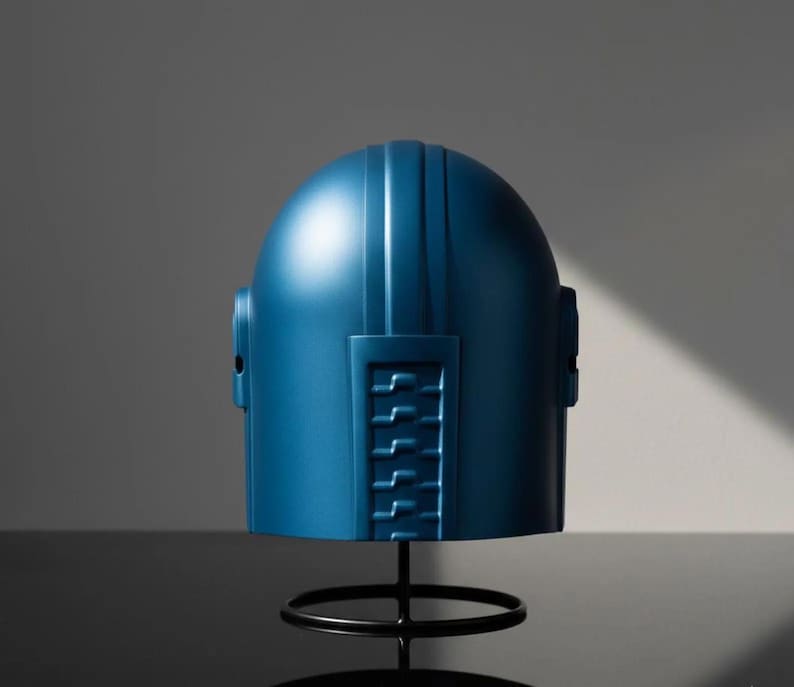 Puede incluir: Un casco azul con una parte superior redondeada y l&iacute;neas verticales, montado sobre un soporte de metal negro. El casco tiene un dise&ntilde;o rectangular en el lateral con una serie de l&iacute;neas horizontales. El fondo es gris neutro.