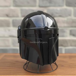 Op de afbeelding: Een glanzende zwarte helm met een opvallend vizier en een T-vormige opening. De helm wordt getoond op een draadstandaard, met de tekst "medievalforgeco." zichtbaar aan de zijkant. Het ontwerp van de helm suggereert een futuristisch of sciencefiction thema.