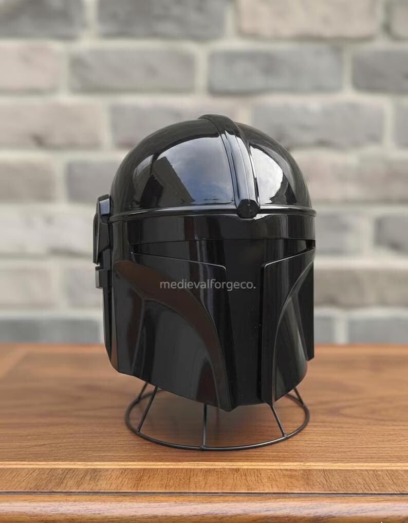 Op de afbeelding: Een glanzende zwarte helm met een opvallend vizier en een T-vormige opening. De helm wordt getoond op een draadstandaard, met de tekst "medievalforgeco." zichtbaar aan de zijkant. Het ontwerp van de helm suggereert een futuristisch of sciencefiction thema.