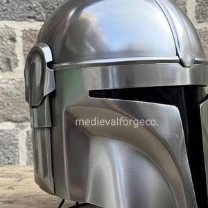 Op de afbeelding: Een close-up van een metallic zilveren helm met een vizier en een afgeronde bovenkant. De helm heeft een gladde, gepolijste afwerking en een donker interieur. De tekst "medievalforgeco." is zichtbaar op de zijkant. De helm is waarschijnlijk een kostuum of verzamelobject.