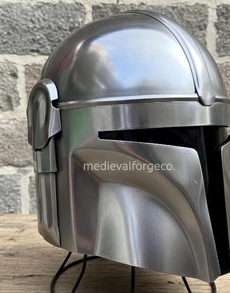 Op de afbeelding: Een close-up van een metallic zilveren helm met een vizier en een afgeronde bovenkant. De helm heeft een gladde, gepolijste afwerking en een donker interieur. De tekst "medievalforgeco." is zichtbaar op de zijkant. De helm is waarschijnlijk een kostuum of verzamelobject.