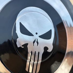 May include: A circular, metallic object with a black center featuring a silver skull emblem. The skull has a distinctive design with sharp angles and vertical lines. The outer rim is silver, creating a contrasting border.