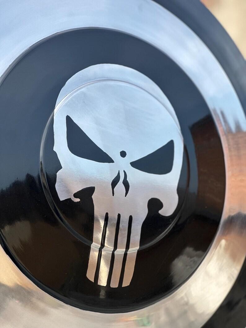May include: A circular, metallic object with a black center featuring a silver skull emblem. The skull has a distinctive design with sharp angles and vertical lines. The outer rim is silver, creating a contrasting border.