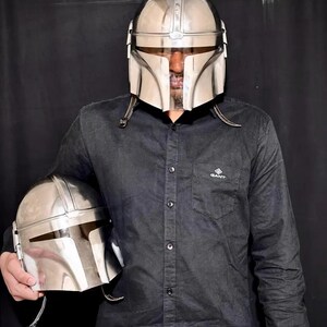 Może przedstawiać: Osoba nosi błyszczący srebrny hełm Mandalorianina, z drugim hełmem w dłoni. Hełmy mają charakterystyczny wizjer w kształcie litery T i są wykonane z odblaskowego materiału. Osoba ma na sobie czarną koszulę zapinaną na guziki.