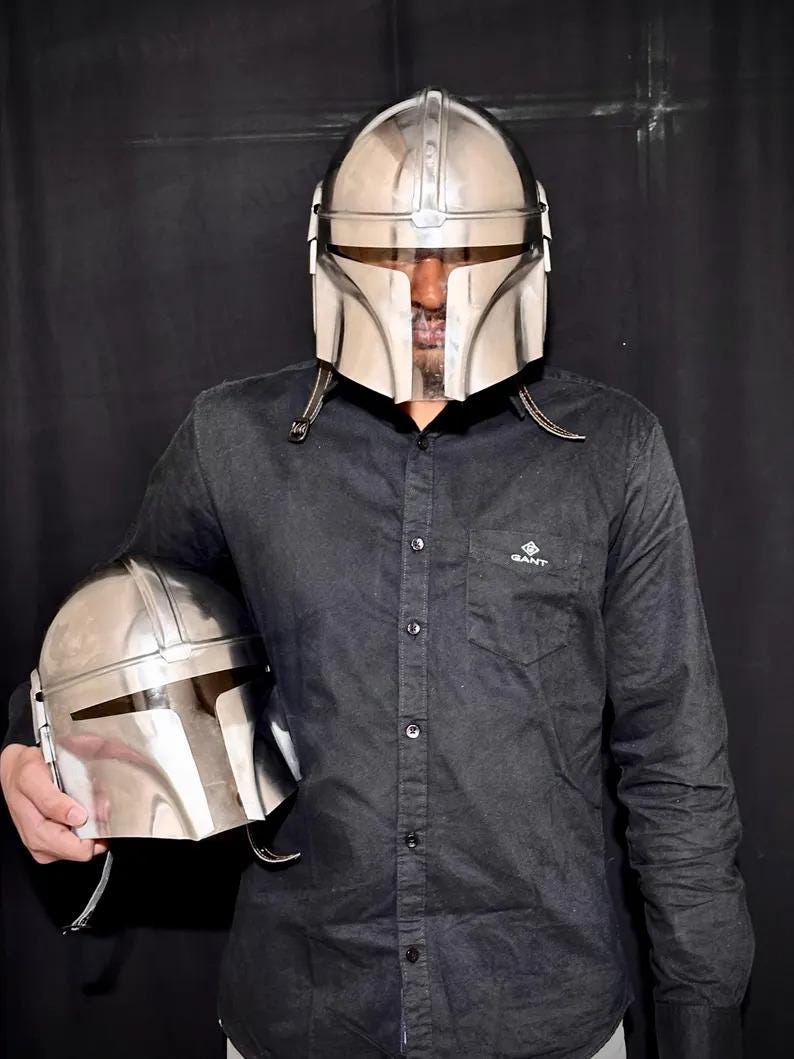 Może przedstawiać: Osoba nosi błyszczący srebrny hełm Mandalorianina, z drugim hełmem w dłoni. Hełmy mają charakterystyczny wizjer w kształcie litery T i są wykonane z odblaskowego materiału. Osoba ma na sobie czarną koszulę zapinaną na guziki.