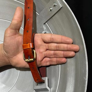 May include: A silver-colored shield with a brown leather strap and a gold-colored buckle. The shield has a circular design with concentric lines. The strap is attached to the back of the shield with metal brackets.
