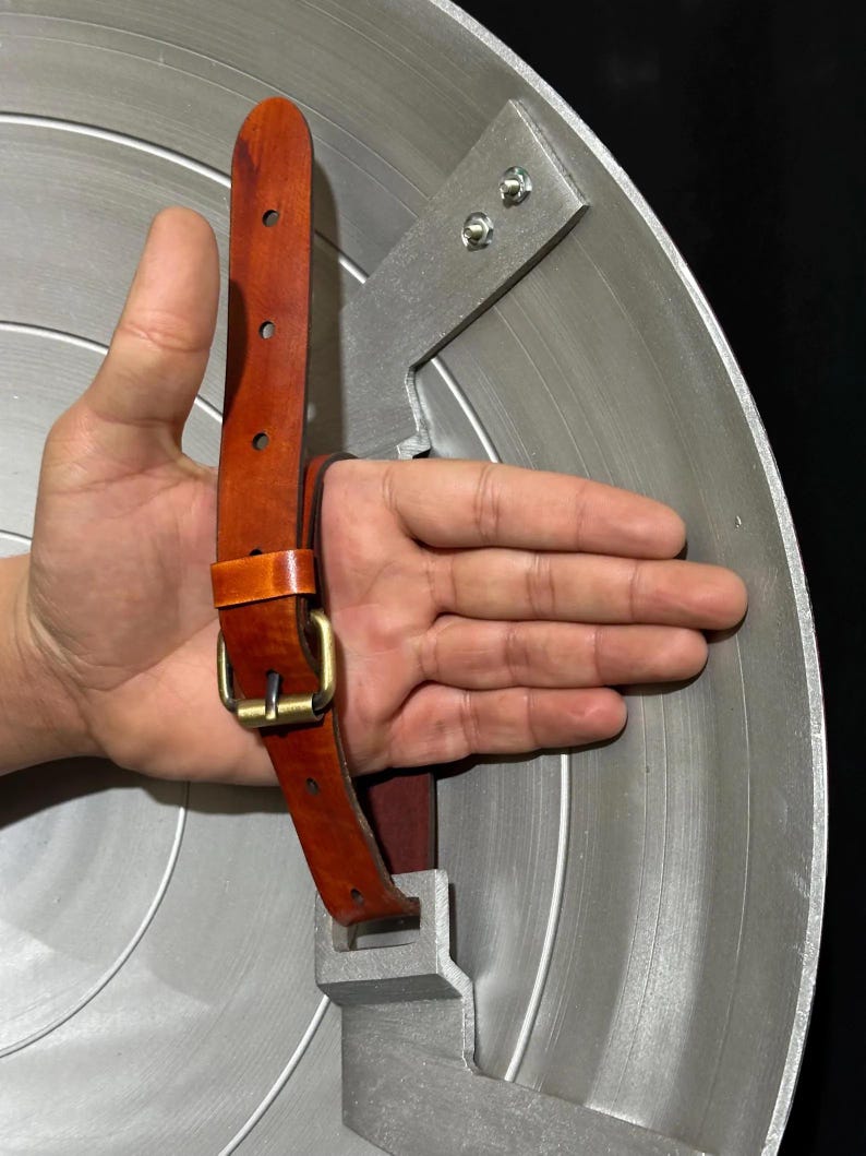 May include: A silver-colored shield with a brown leather strap and a gold-colored buckle. The shield has a circular design with concentric lines. The strap is attached to the back of the shield with metal brackets.