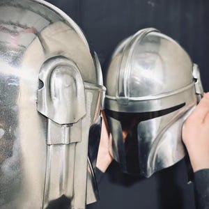 Może przedstawiać: Dwa srebrne hełmy o metalicznym połysku. Hełmy mają zaokrąglony kształt ze szczeg&oacute;łowymi grzbietami i wizjerami. Jeden hełm jest trzymany przez osobę, a drugi znajduje się na pierwszym planie. Tło jest ciemne.