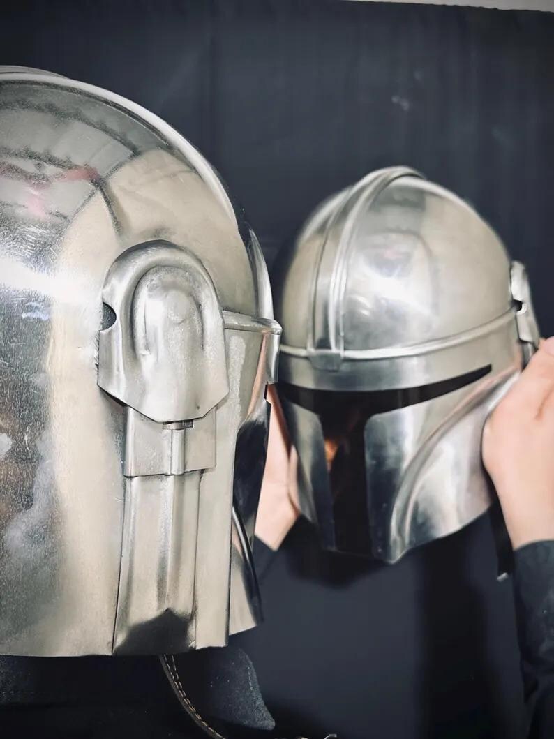 Może przedstawiać: Dwa srebrne hełmy o metalicznym połysku. Hełmy mają zaokrąglony kształt ze szczeg&oacute;łowymi grzbietami i wizjerami. Jeden hełm jest trzymany przez osobę, a drugi znajduje się na pierwszym planie. Tło jest ciemne.