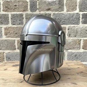 Op de afbeelding: Een zilverkleurige helm met een zwart vizier, gepresenteerd op een zwarte draadstandaard. De helm heeft een afgeronde bovenkant en hoekige kenmerken, met de tekst "...9e10t16veib9m" zichtbaar aan de zijkant. De achtergrond is een bakstenen muur.