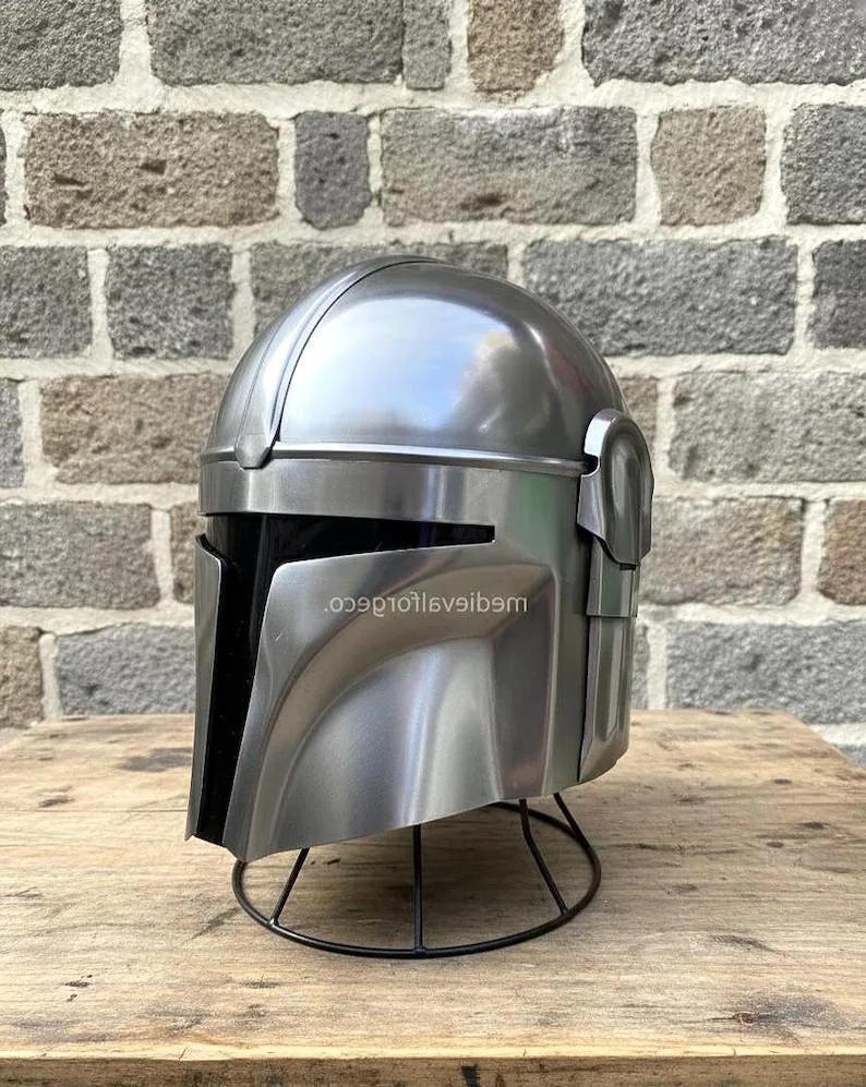 Op de afbeelding: Een zilverkleurige helm met een zwart vizier, gepresenteerd op een zwarte draadstandaard. De helm heeft een afgeronde bovenkant en hoekige kenmerken, met de tekst "...9e10t16veib9m" zichtbaar aan de zijkant. De achtergrond is een bakstenen muur.