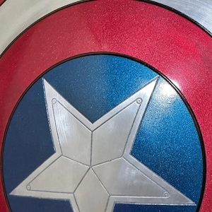May include: Close-up of a Captain America shield. The shield features a silver star on a blue circle, surrounded by a red ring and a silver outer rim. The design is a classic superhero emblem.