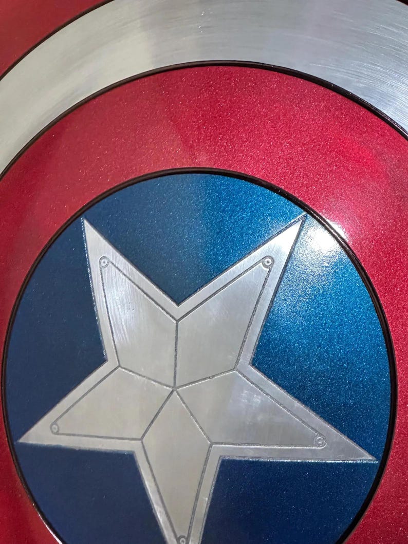 May include: Close-up of a Captain America shield. The shield features a silver star on a blue circle, surrounded by a red ring and a silver outer rim. The design is a classic superhero emblem.